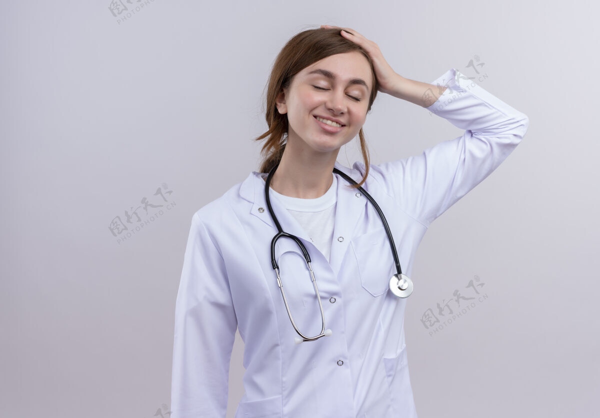 女人面带微笑的年轻女医生 穿着医用长袍 戴着听诊器 将手放在头上 放在隔离的白色墙壁上 留有复印空间年轻医生穿