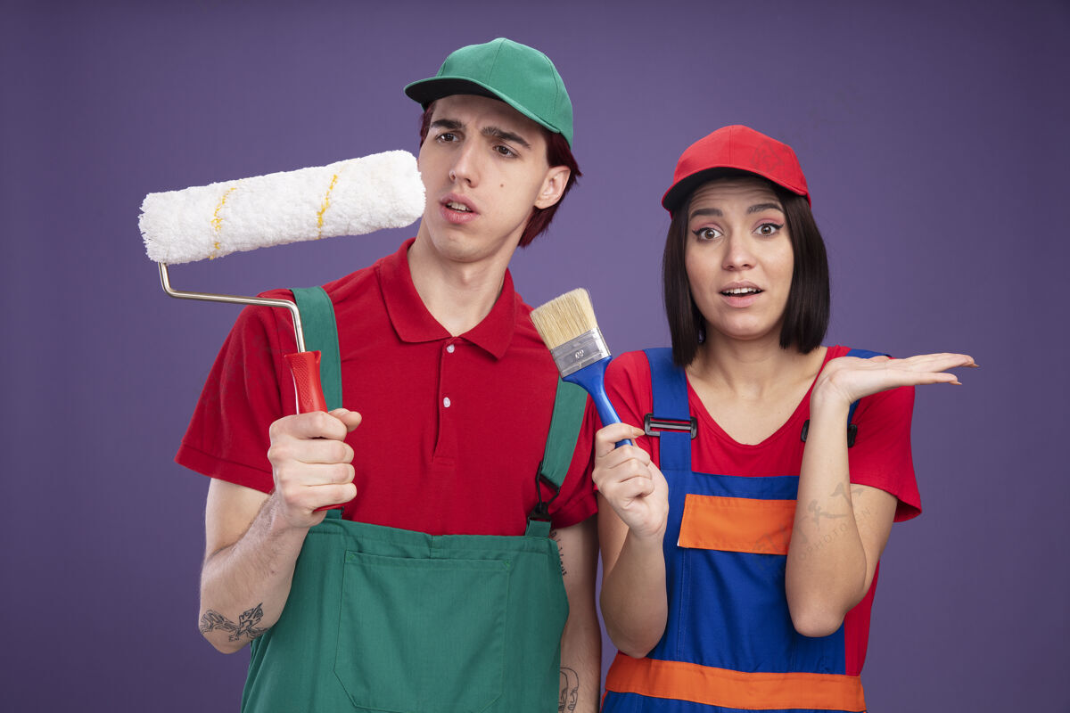 帽子一对穿着建筑工人制服 戴着帽子的年轻夫妇困惑地看着手里拿着油漆辊的男孩 印象深刻的女孩手里拿着油漆刷 在紫色的墙上露出空手女孩困惑印象