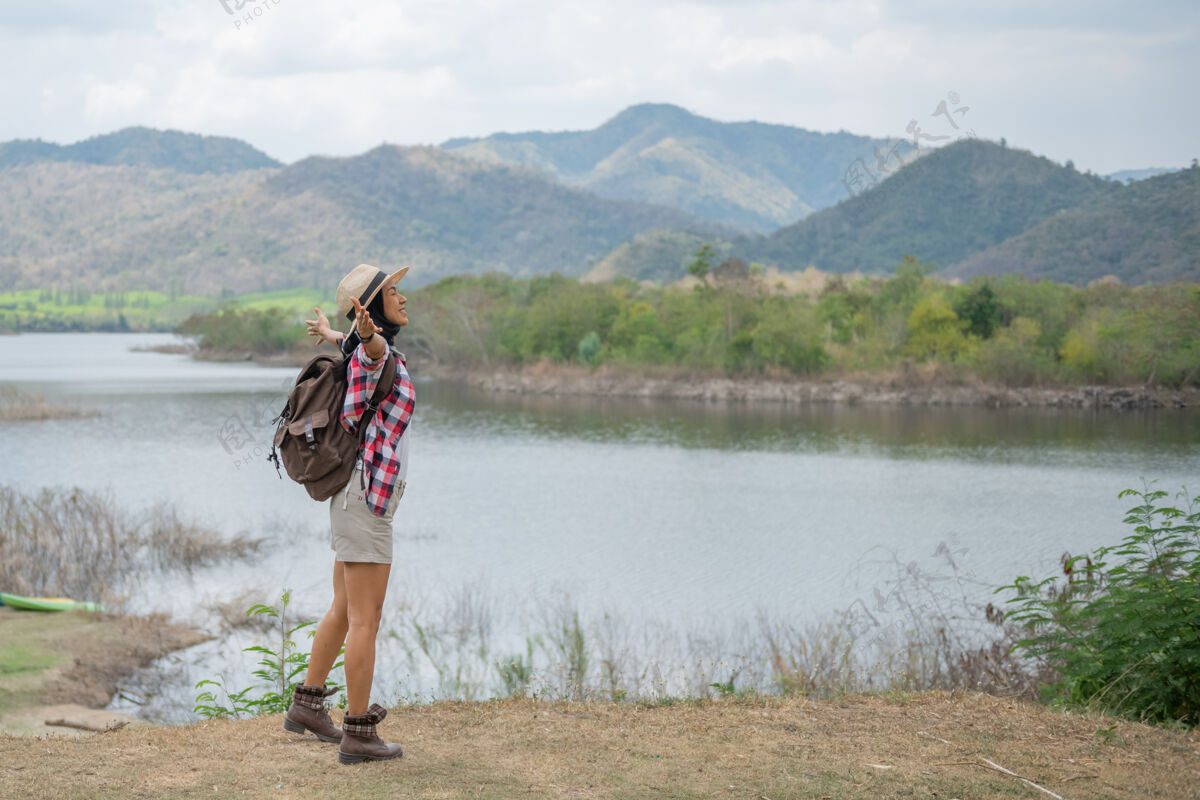 露营在湖畔举手的女士\/前面的亚洲女徒步旅行者微笑着快乐 在树林里徒步旅行的女士 温暖的夏日活动年轻女性