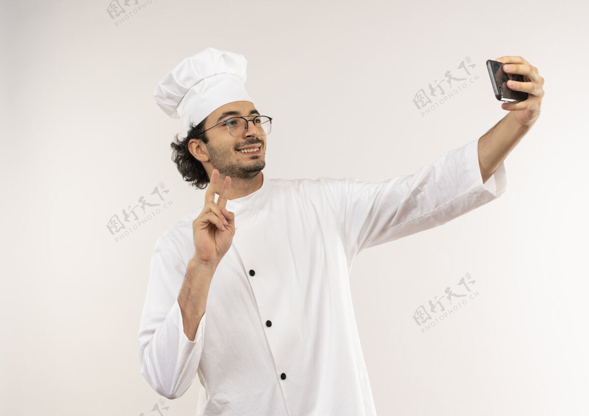 年轻面带微笑的年轻男厨师身着厨师制服 戴着眼镜自拍 在白墙上显示出与世隔绝的和平姿态手势男性采取