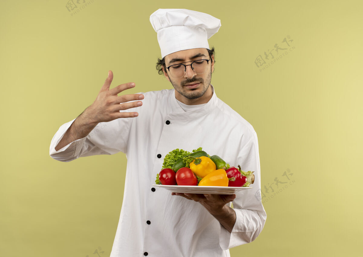 男性年轻的男厨师闭着眼睛 穿着厨师制服 戴着眼镜 拿着和嗅着隔离在绿墙上的盘子里的蔬菜眼镜烹饪封闭