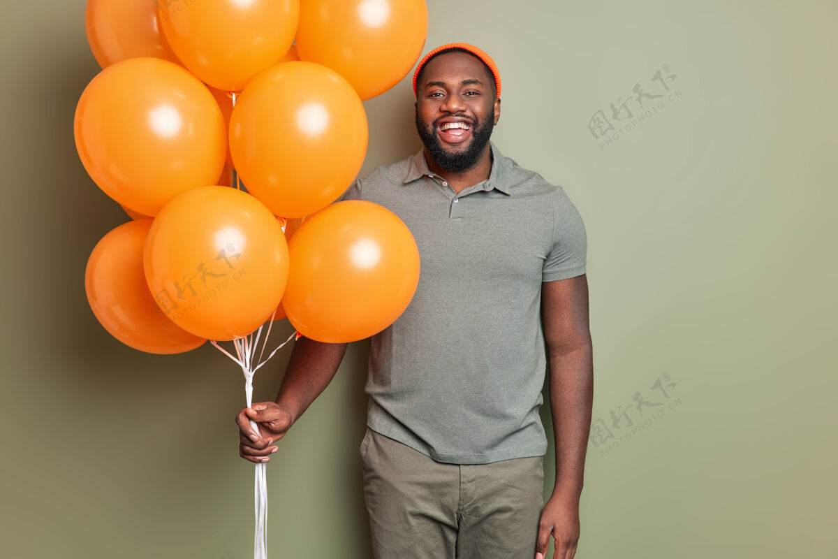男人快乐的男人站在那里 高兴地穿着休闲服 手里拿着一堆橙色充气气球 对着卡其色的墙在室内摆姿势氦卡其色帅气