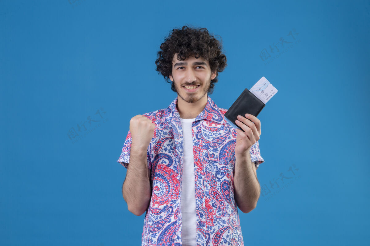 年轻面带微笑的年轻英俊的旅行家男子举着钱包和机票 高举拳头站在与世隔绝的蓝色墙壁上 留着复印空间钱包举起来机票