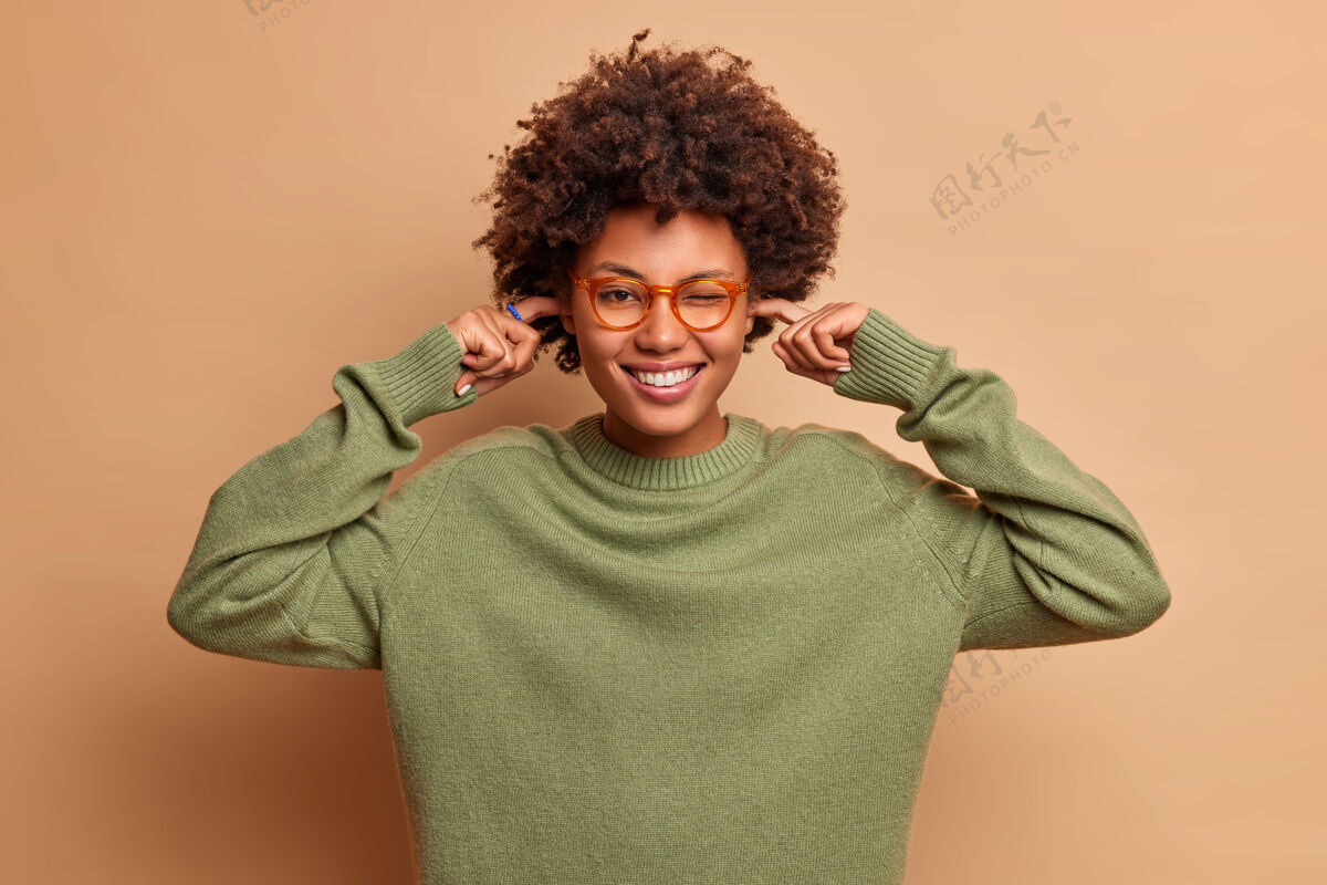 肖像积极的卷发女人开心地笑着塞住耳朵避免很大声的音乐戴着透明的眼镜和毛衣有快乐的心情隔着棕色的墙套头衫休闲高兴