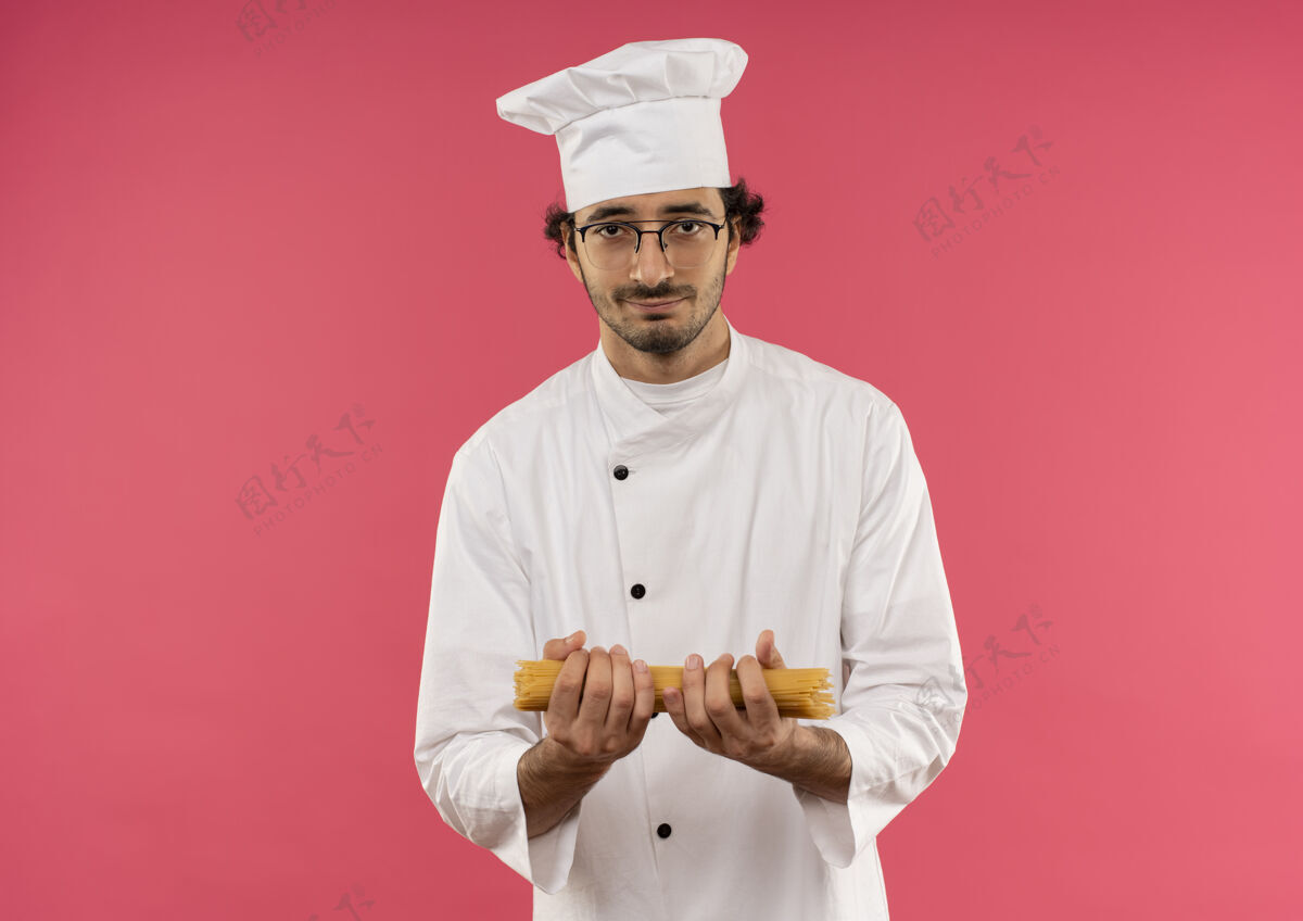 请穿年轻的男厨师穿着厨师制服 戴着眼镜 把意大利面孤立地放在粉红色的墙上意大利面眼镜年轻
