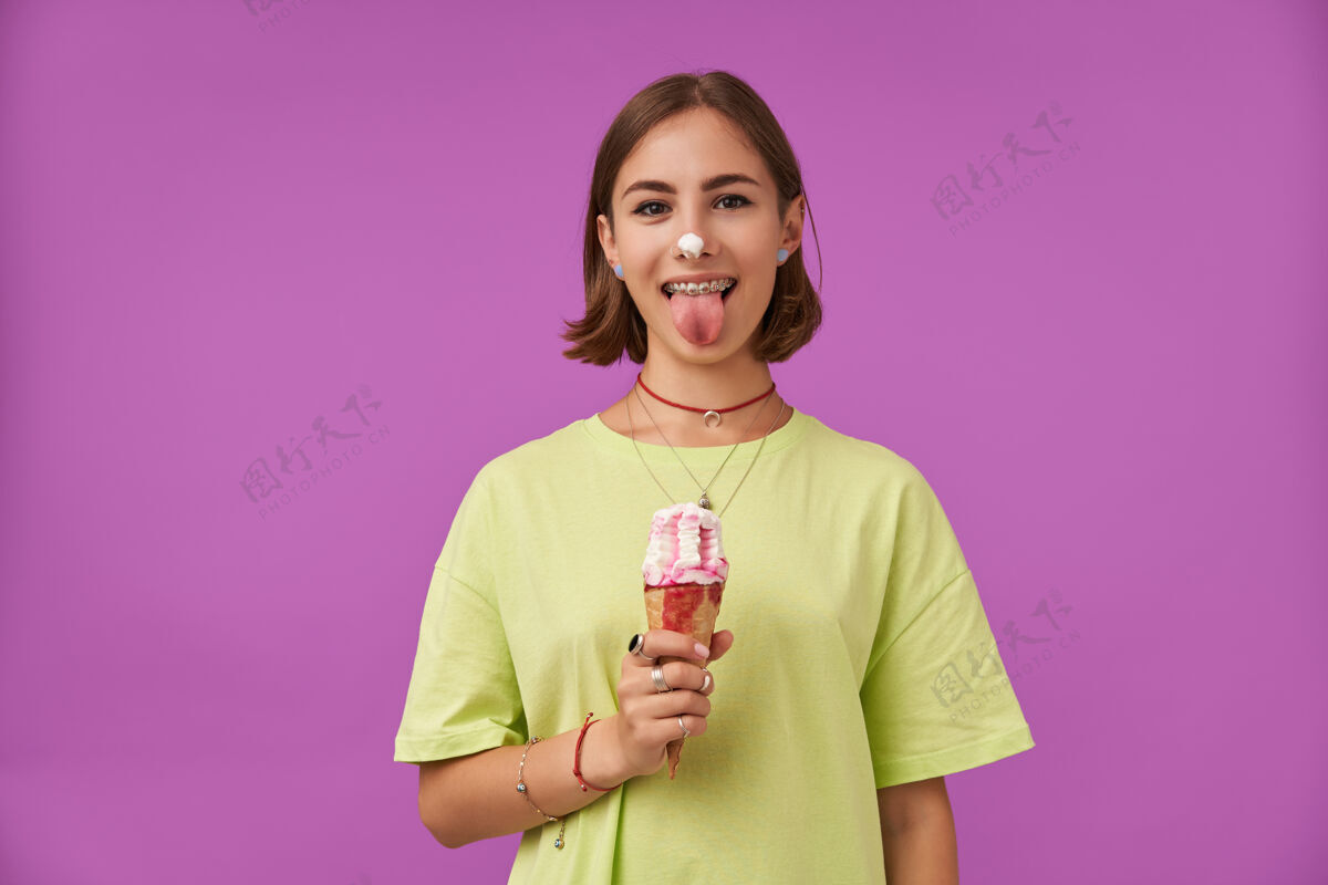 项链女学生 年轻的女士拿着冰淇淋 露出舌头冰淇淋放在鼻子上站在紫色的墙上穿着绿色的t恤 牙套 戒指 项链和手镯牙套独自青少年