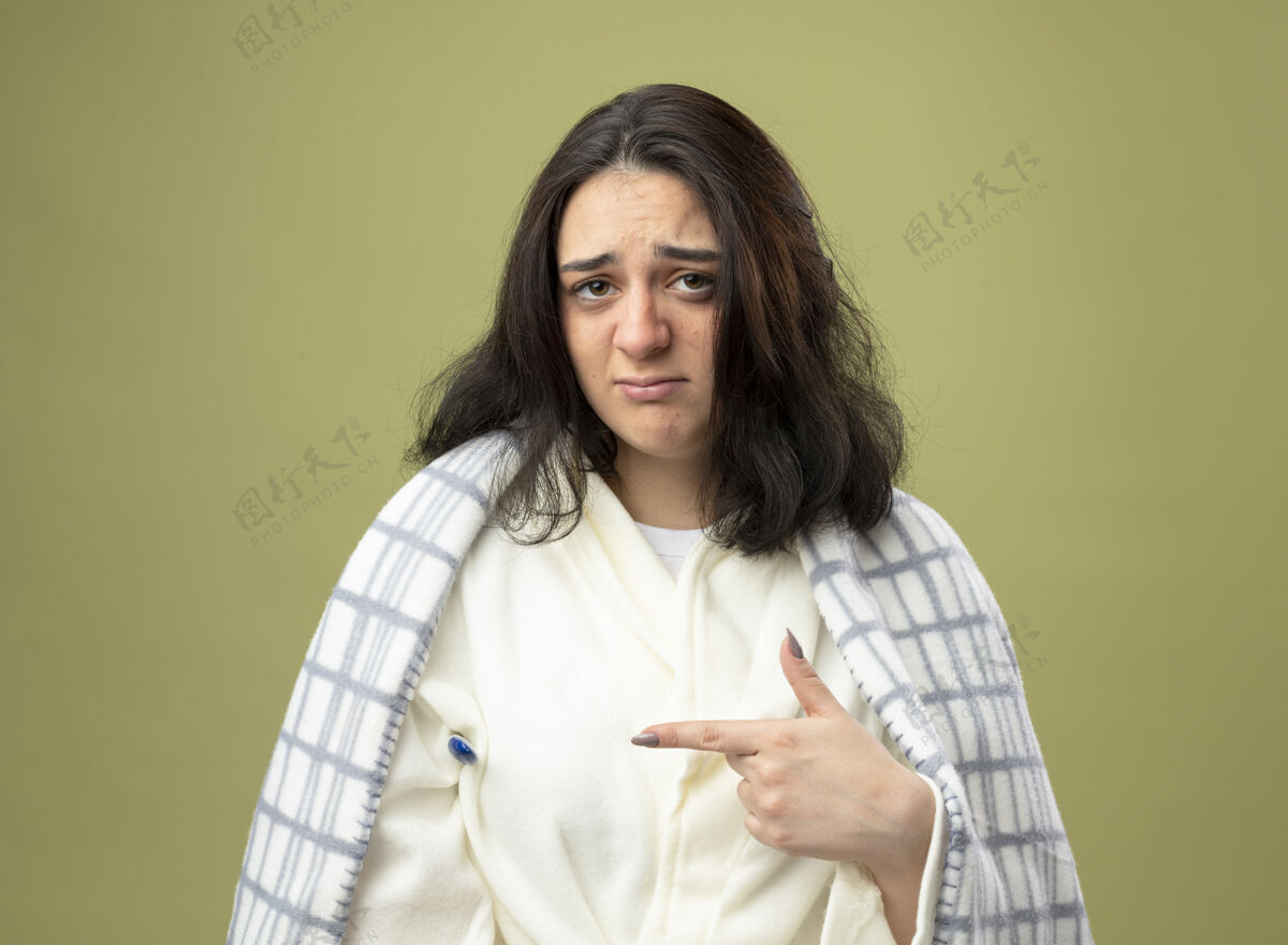 格子弱小的年轻白种人生病的女孩穿着格子布裹着的长袍 看着摄像机 腋下拿着温度计 指着橄榄绿背景上孤立的温度计疾病年轻橄榄