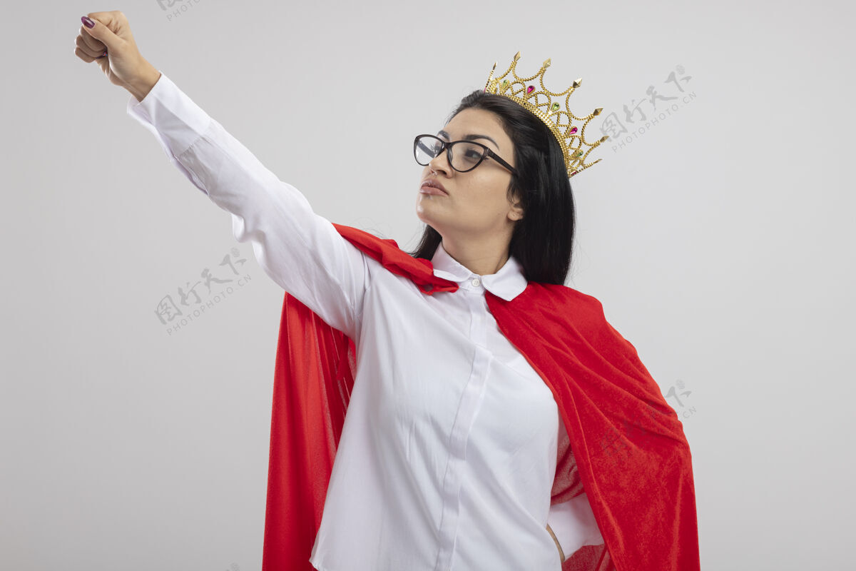 眼镜自信的年轻白种人超级英雄女孩戴着眼镜 戴着王冠 手放在腰上 举起拳头 看着她在白色背景下孤立的拳头超级英雄手拳头