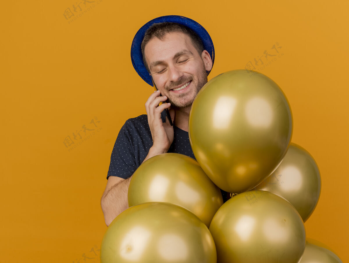 氦微笑英俊的白人男子戴着蓝色的派对帽手持氦气球在电话上交谈 橙色背景上有复印空间气球派对电话