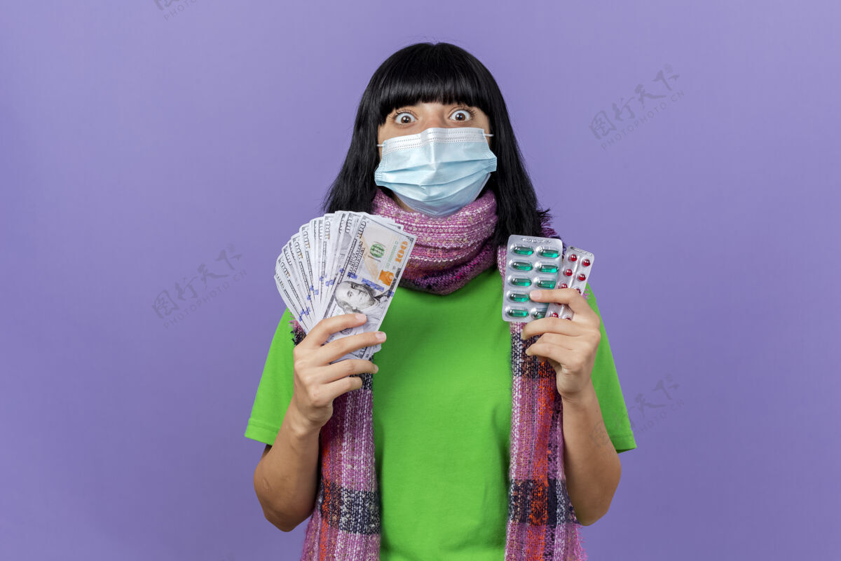钱印象深刻的年轻病高加索女孩戴着面具和围巾拿着钱和胶囊包看着相机隔离在紫色背景与复制空间包印象疾病
