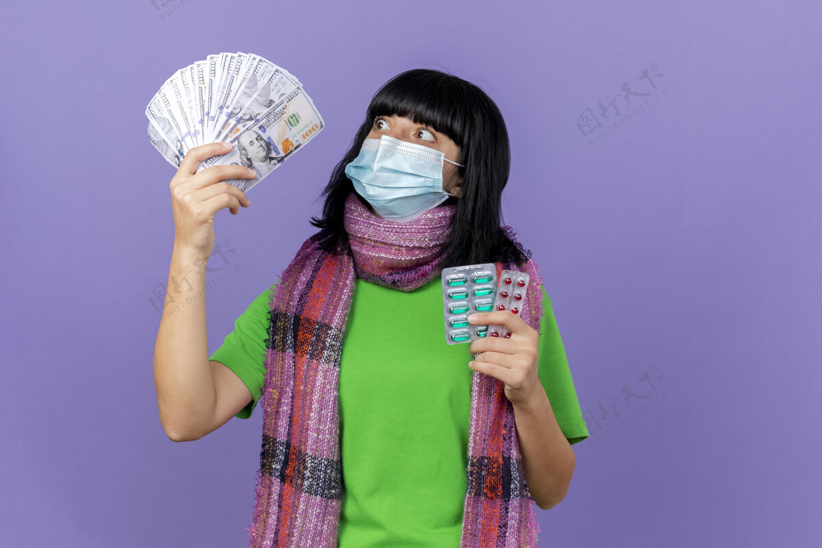 包装印象深刻的年轻病高加索女孩戴着面具和围巾拿着钱和胶囊包看着钱孤立的紫色背景与复制空间抱疾病女孩