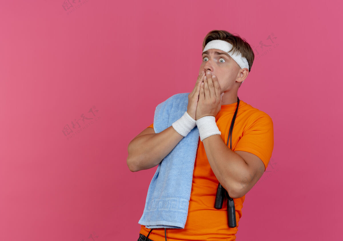 运动焦急的年轻帅气的运动型男子戴着头带和腕带 用毛巾和跳绳围住脖子 看着笔直的手放在嘴上 隔离在粉色的空间里复制年轻毛巾