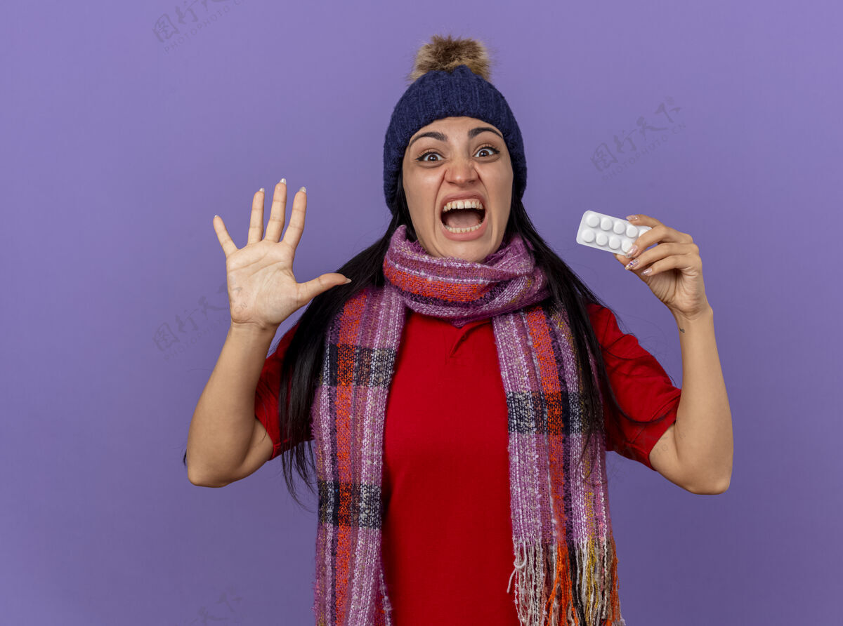 女孩令人印象深刻的年轻白种人生病的女孩戴着冬天的帽子和围巾 展示一包平板电脑 看着相机 显示五只手孤立在紫色背景上药片印象包