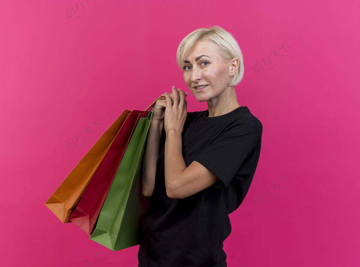 相机微笑的中年金发斯拉夫妇女站在侧视图拿着购物袋看着摄像机在深红的背景与复制空间隔离微笑购物金发
