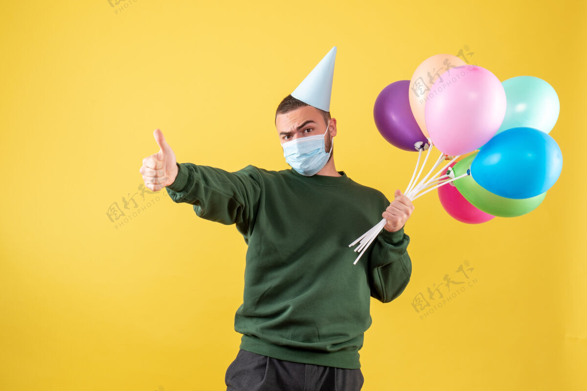流行正面图：黄色背景上戴着面具手持彩色气球的年轻男性公园成人年轻男性