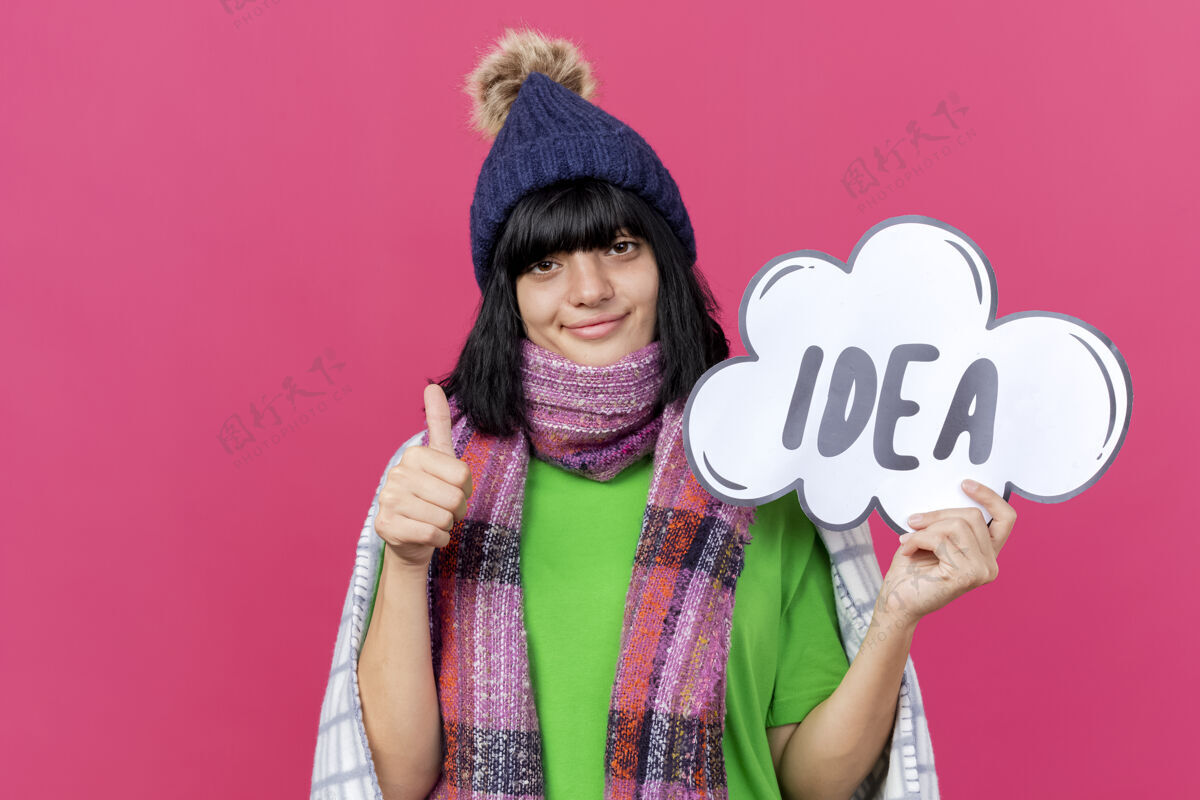 创意高兴的年轻病高加索女孩戴着冬季帽子和围巾包裹在格子布举行的想法泡泡看相机显示拇指向上红色背景与复制空间孤立抱围巾拇指