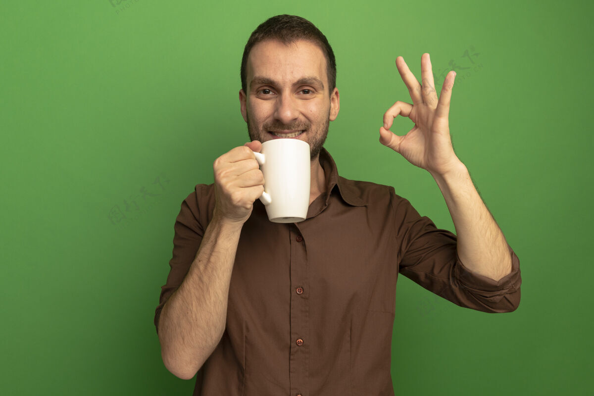 微笑微笑着的年轻白人男子端着一杯茶看着相机做着ok标志 隔离在绿色背景上 留着复印空间拿着年轻杯子
