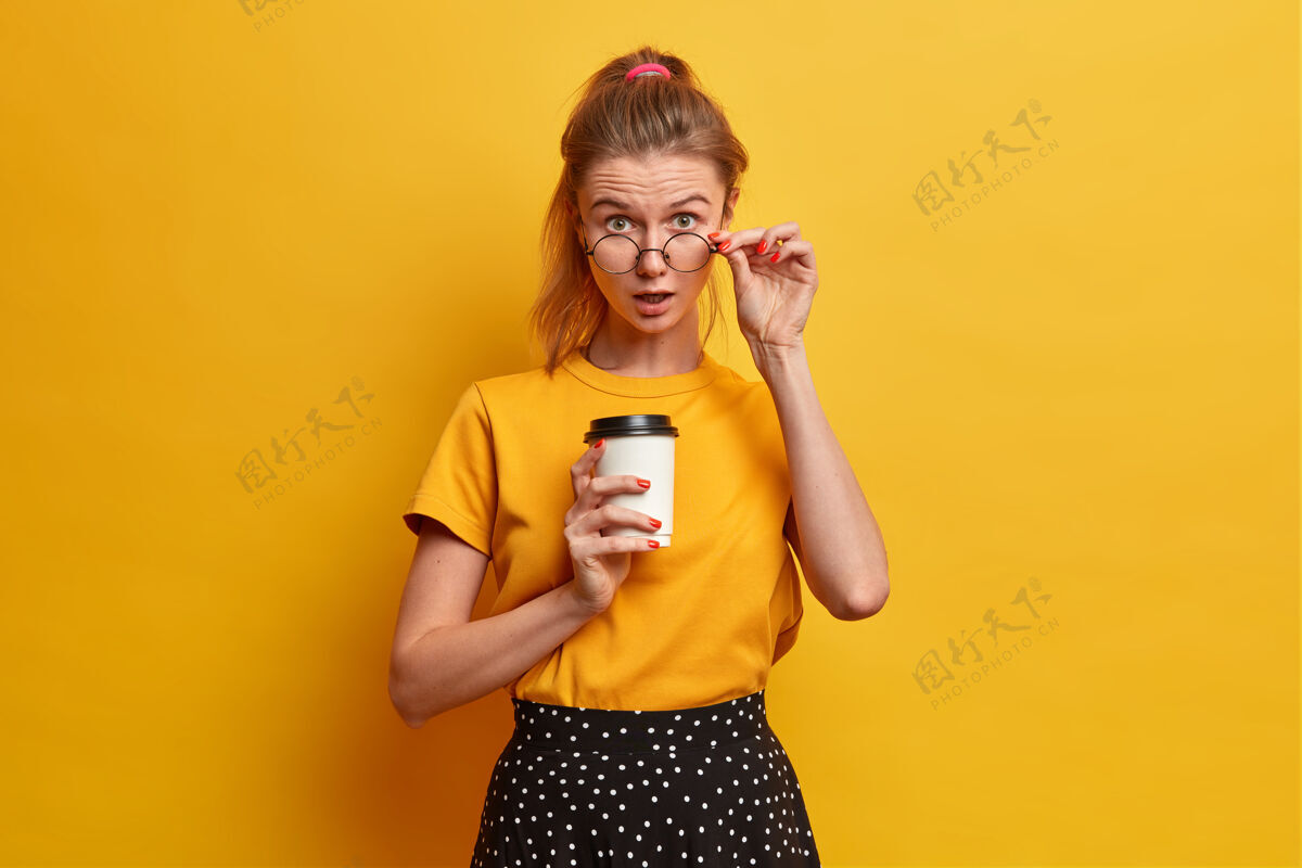 女性惊诧的欧洲女学生透过眼镜看 拿着外卖咖啡摆姿势 听到惊诧的消息 不敢相信自己的眼睛 穿着休闲服印象人水平