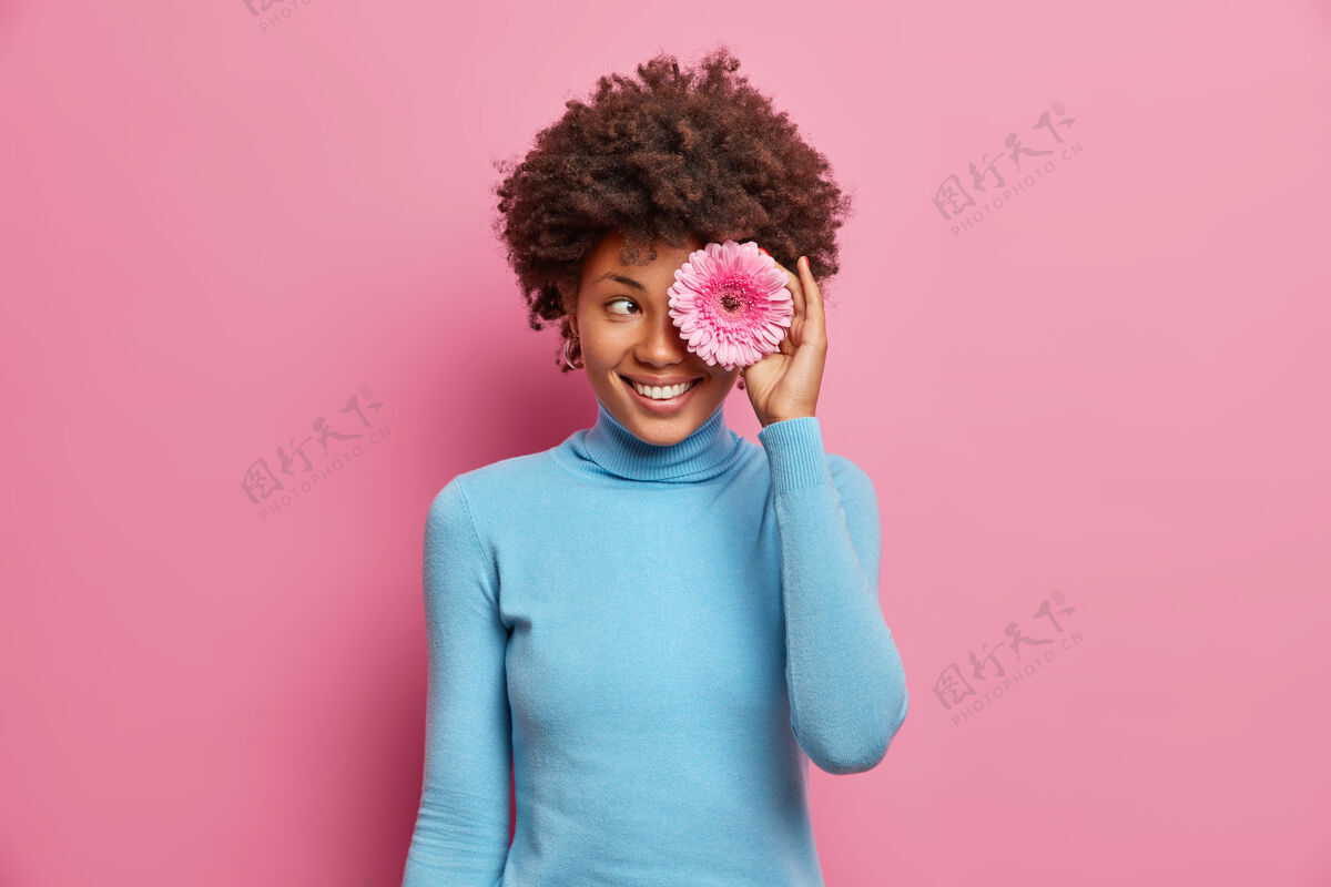 美国快乐积极的黑皮肤年轻女子用粉色非洲菊遮住眼睛 露出牙齿般的微笑 穿着随意 在室内摆姿势 享受春天的时光花卷曲微笑