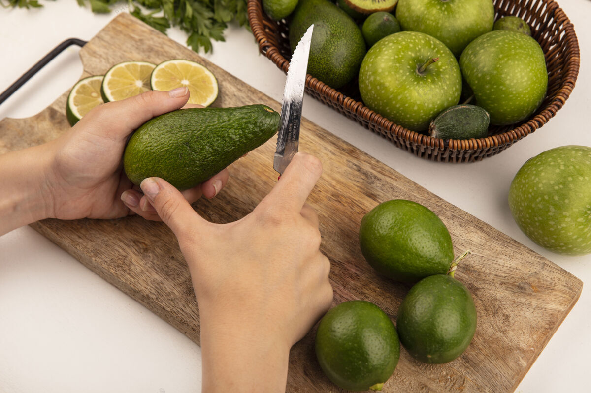 刀女性手用刀在木制厨房板上切新鲜鳄梨的俯视图 白色墙上的桶上放着酸橙和苹果视图美食女