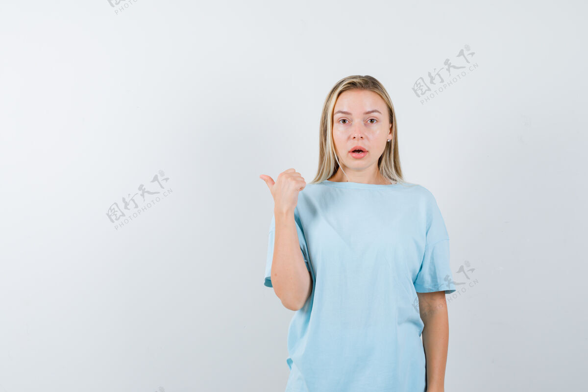 情感穿着蓝色t恤的金发女孩用拇指指着一边 看上去很惊讶 正前方视图拇指夏天大海