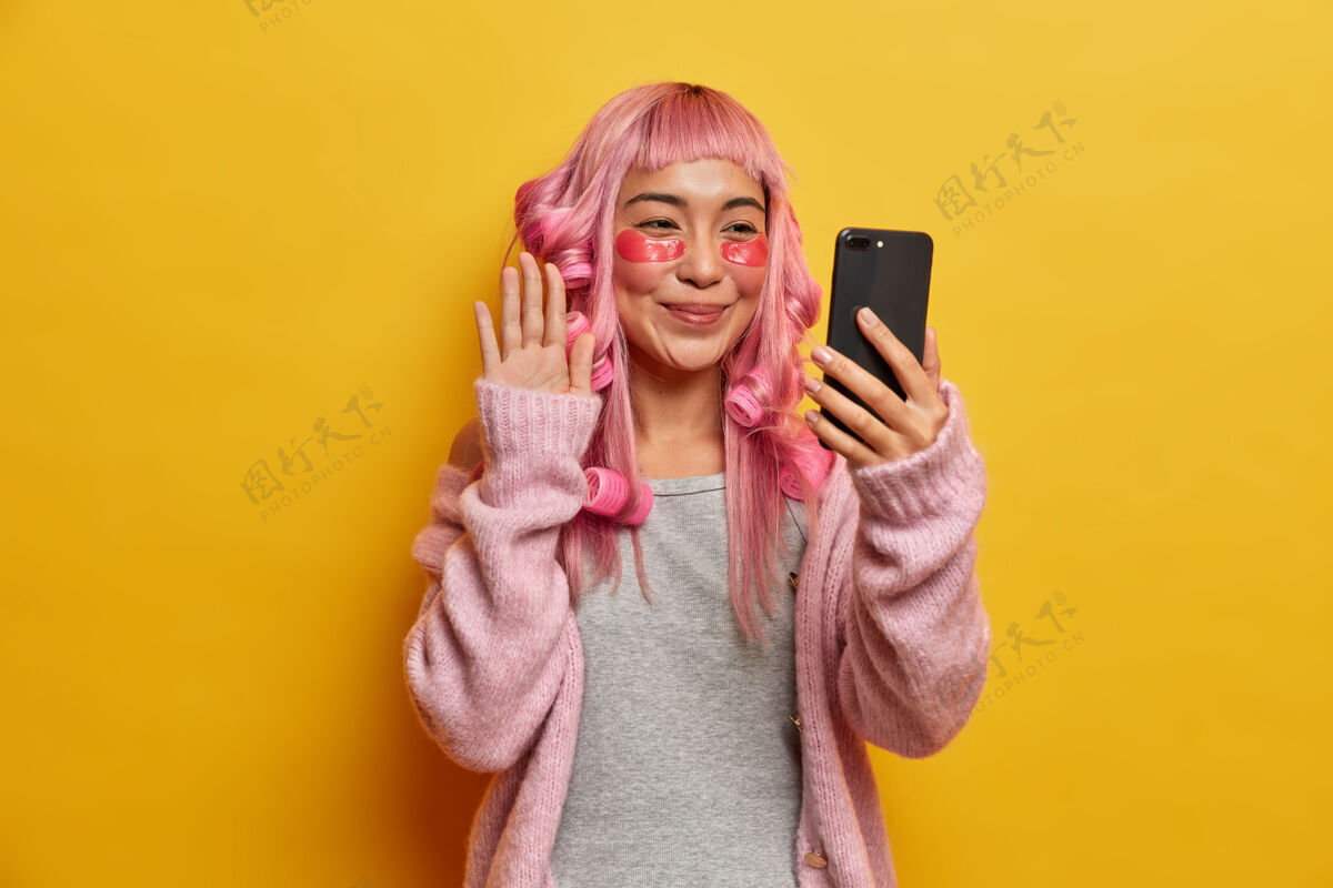 内容积极美丽的年轻女子接受美容程序 在染成粉红色的头发上戴上滚轴 在眼睛下涂抹胶原蛋白垫 用智能手机自拍请自拍美丽