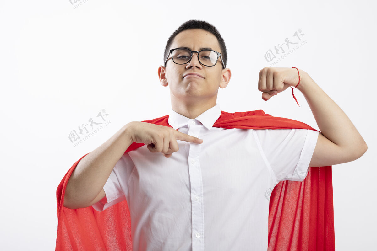 白色穿着红斗篷的自信的年轻超级英雄男孩戴着眼镜做着强有力的手势看着摄像机指着他隔离在白色背景上的肌肉男孩穿着自信