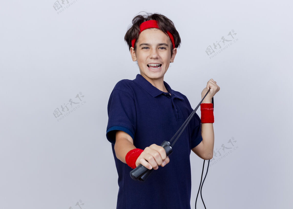 运动快乐的年轻帅气的运动男孩戴着头带和戴着牙套的腕带看着相机拉着跳绳在白色背景上与复制空间隔离背带拉腕带