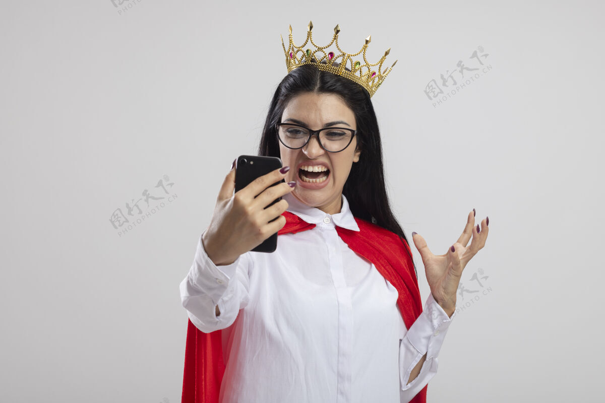 女孩愤怒的年轻白种人超级英雄女孩戴着眼镜 戴着王冠 手举着手机看着 在白色的背景上与外界隔绝 留着复印空间眼镜佩戴白色