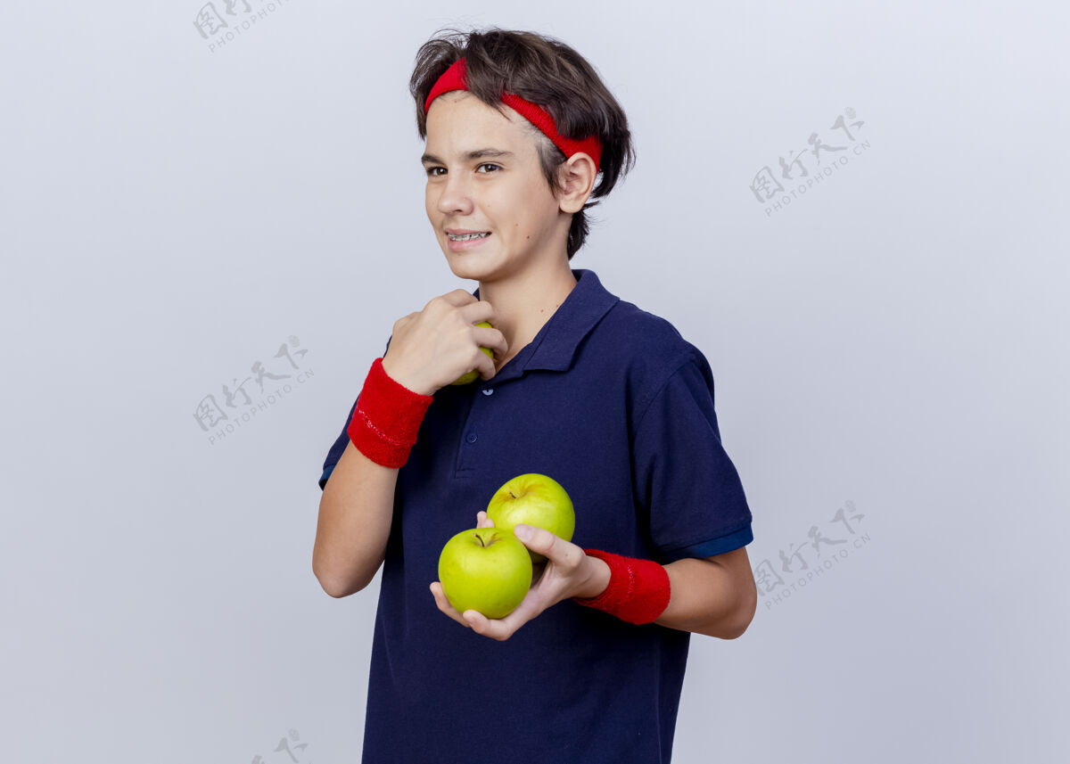 持有面带微笑的年轻帅气的运动男孩戴着头带和护腕 戴着牙套 看起来笔直地拿着苹果 孤立地放在白色背景上 留着复印空间腕带头带运动