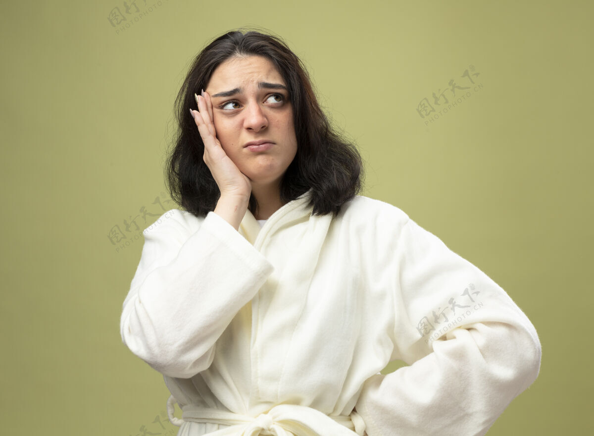 幼焦急的年轻白种人生病的女孩穿着长袍 手放在腰上 头贴在橄榄绿的背景上 看着孤立的一面腰青壁板