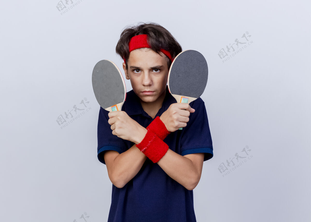 年轻自信的年轻帅气的运动男孩戴着头带和护腕 戴着牙套 双手交叉 拿着乒乓球拍 看着白色背景上的相机 还有复印空间空间男孩球拍