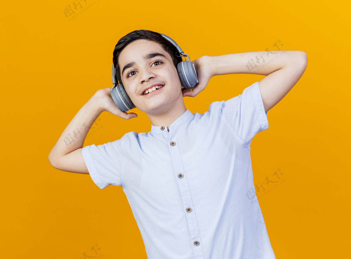 年轻戴着耳机的年轻高加索男孩微笑着抚摸着他们 看着隔离在橙色背景上的摄像机微笑橙色耳机