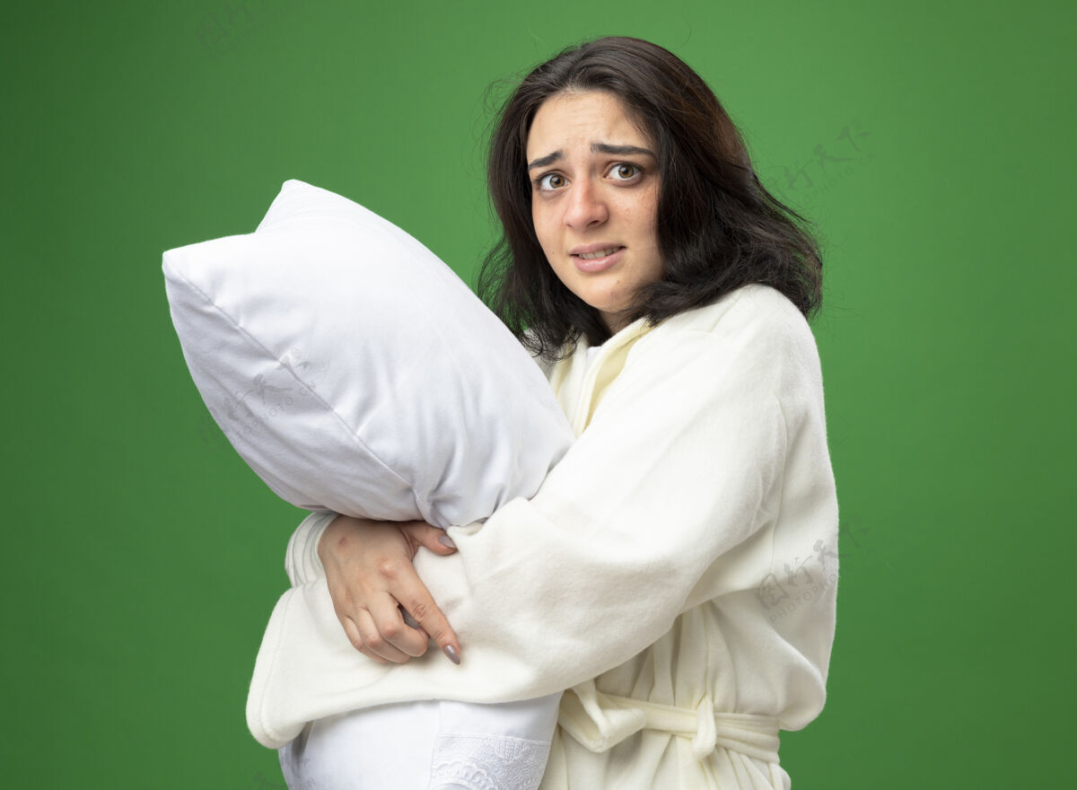 枕头焦急的年轻白种人生病的女孩穿着长袍站在侧视图拥抱枕头看着隔离在绿色背景上的相机女孩视图年轻