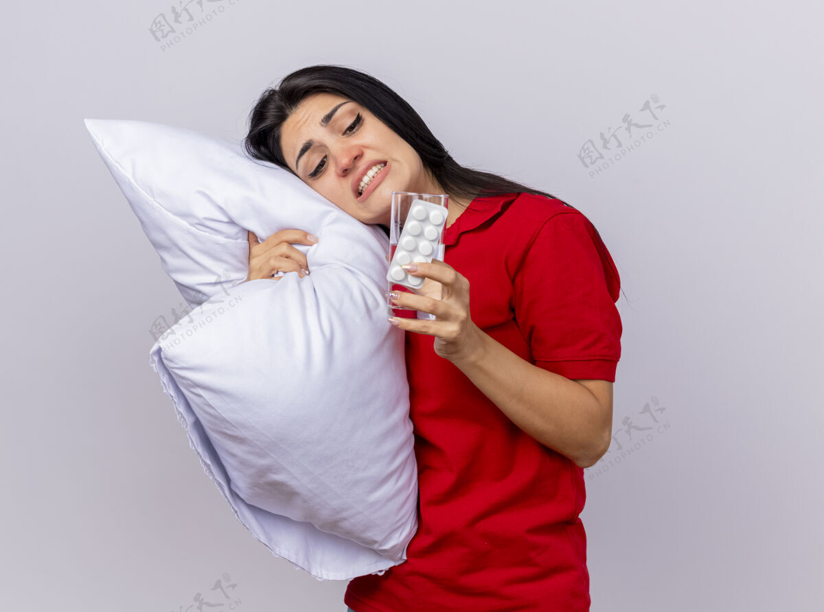 复制恼怒的年轻白人病女孩抱着枕头把头放在枕头上 手里拿着一包药片和一杯水 看着它们孤立地躺在白色背景上 留着复印空间手拥抱年轻