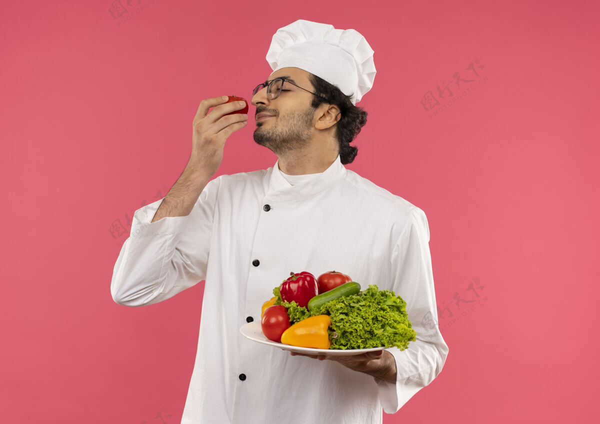 眼镜年轻的男厨师闭上眼睛 高兴地穿着厨师制服 戴着眼镜 手里拿着盘子里的蔬菜 手里还嗅着粉红色的西红柿盘子年轻请