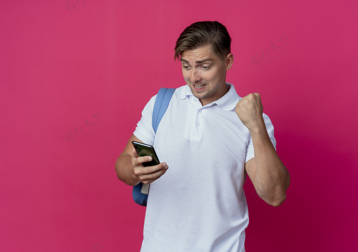 欢乐快乐的年轻帅气的男学生 背着书包 拿着手机 看着手机 在粉红色的墙上显示“是”的手势男人墙秀