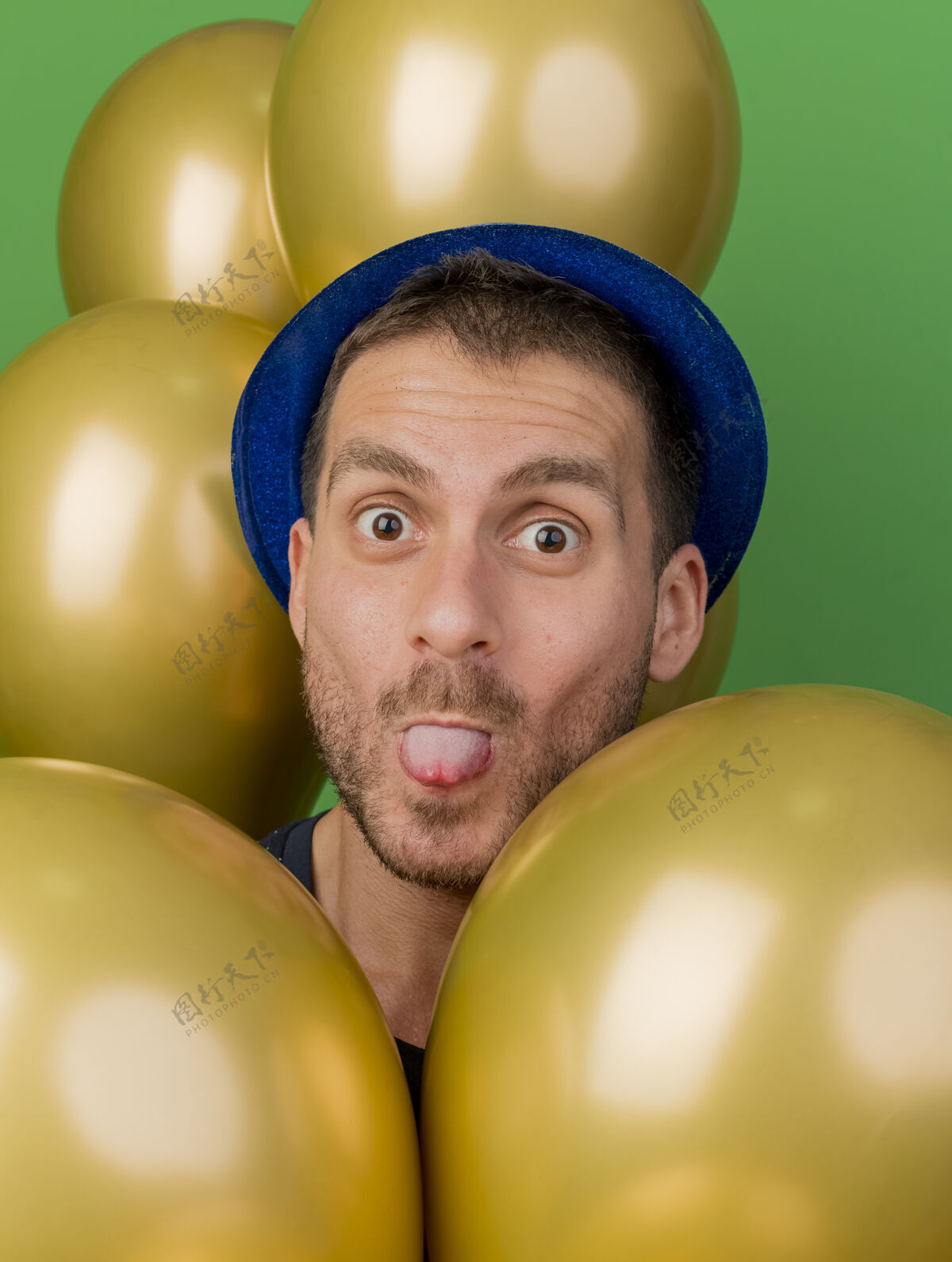 姿势有趣的帅哥戴着蓝色的派对帽 用氦气球在绿色的墙壁上孤立地跺着舌头站着黄金立场人
