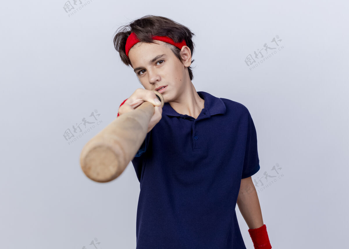 牙齿自信的年轻帅气的运动男孩戴着头带和护腕 戴着牙套看着前面 用棒球棒指着前面 隔离在白色的墙上腕带佩戴站立