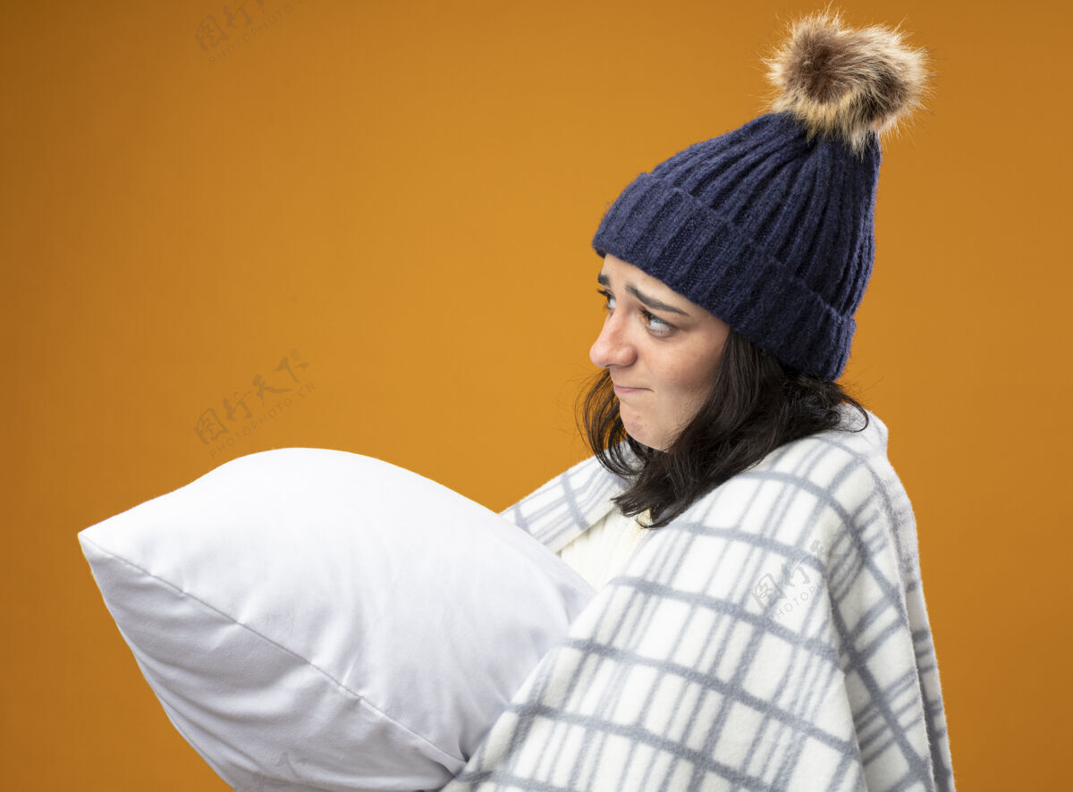 枕头体贴的年轻生病的女人穿着长袍 戴着冬天的帽子 裹着格子呢 站在侧视图里 抱着枕头 在橘色的墙上显得笔直孤立人公民衣服