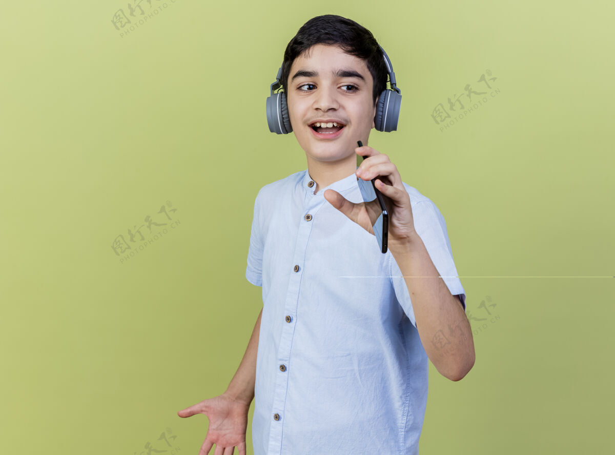 穿着小男孩戴着耳机边唱歌边用手机当麦克风隔离在橄榄绿的墙上人年轻公民