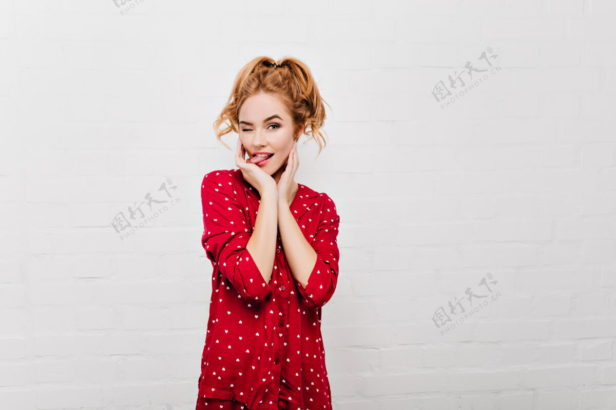 肖像穿着红色睡衣的可爱女士做着滑稽的脸白色墙壁上隔离着穿着睡衣的金发女模特的室内肖像年轻睡眠睡衣