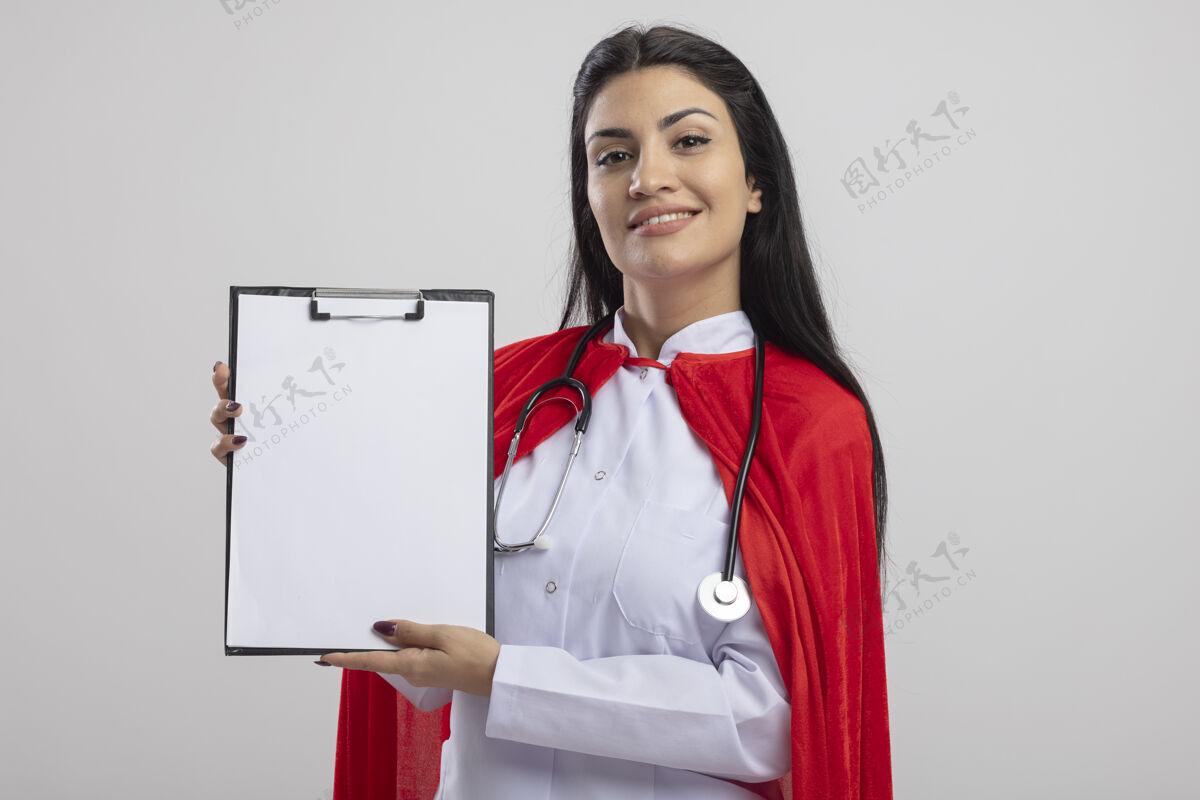 超级英雄微笑的年轻白人超级英雄女孩戴着听诊器显示剪贴板看着相机隔离在白色背景与复制空间剪贴板空间显示