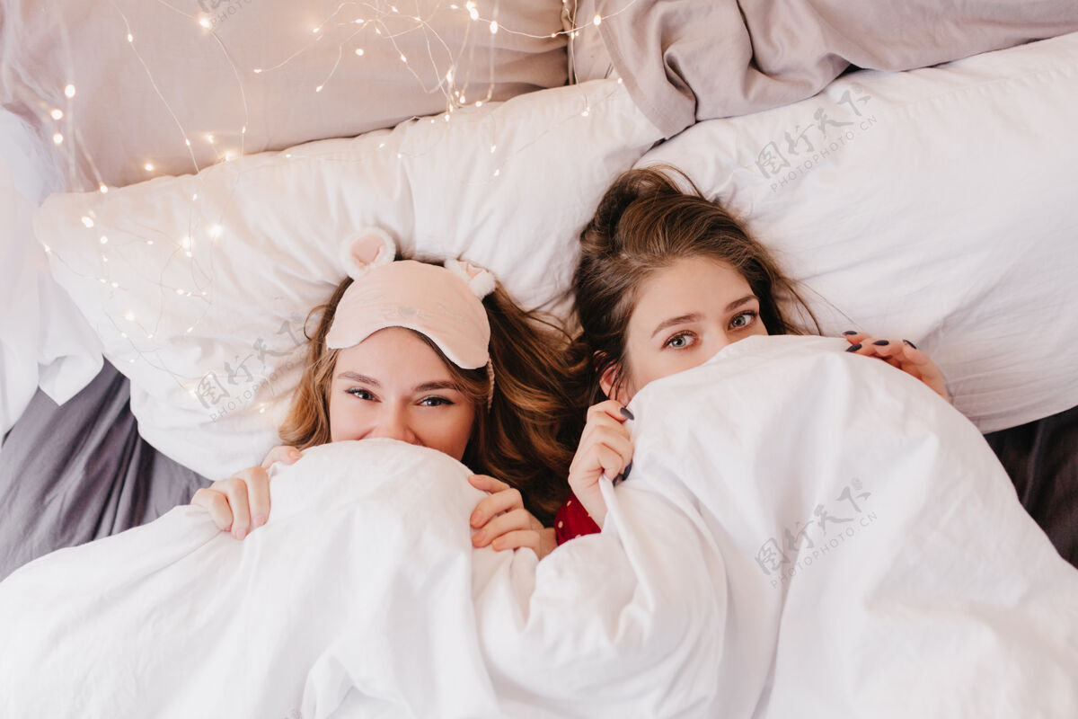 睡眠面罩一大早 兴奋的年轻女士躺在床上 梦幻般的姐妹们躲在毯子下周末成人室内