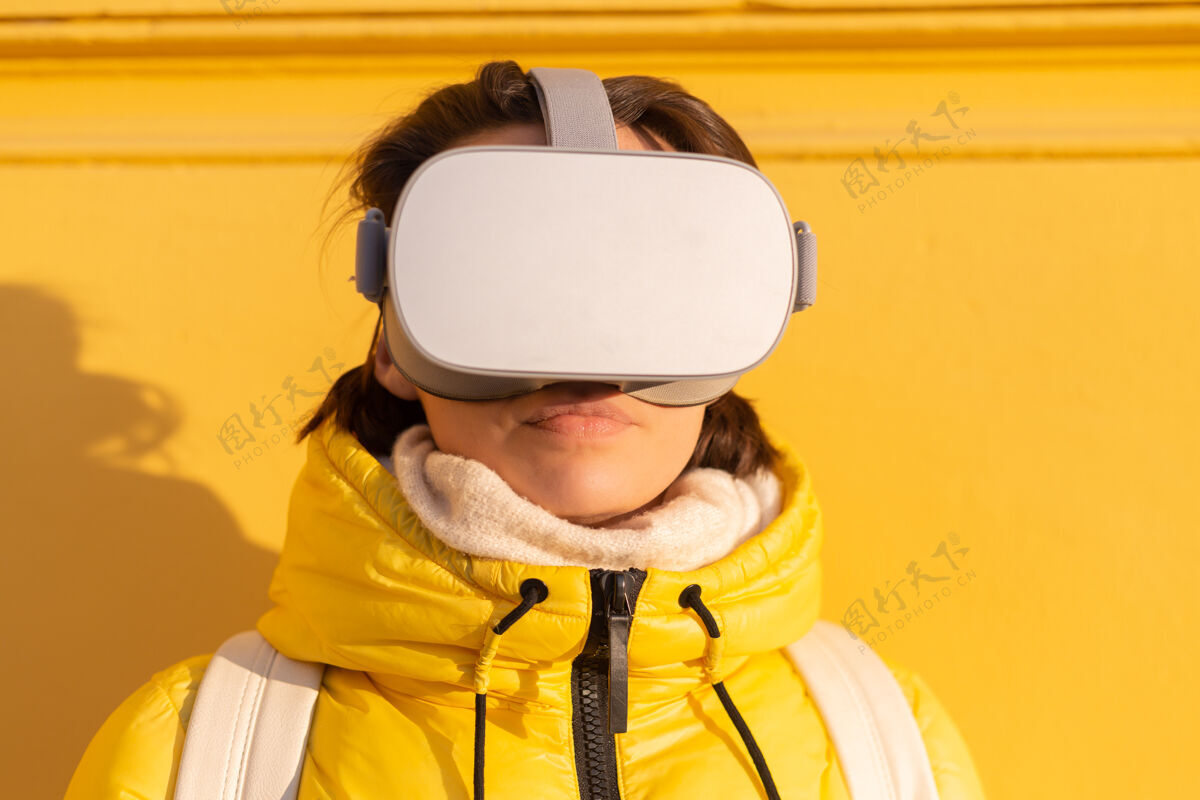可穿戴一个戴着虚拟现实眼镜的女人的肖像 在冬天的阳光下 穿着暖和的衣服 靠在黄色的墙上显示虚拟现实游戏游戏