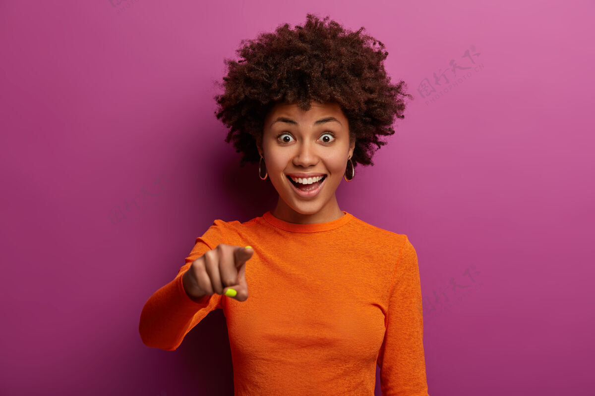 积极快乐的黑皮肤年轻女子的画像用食指笔直地指了指 看到一些惊人的东西 说嘿你 穿着橙色毛衣 隔着充满活力的紫色墙壁反应印象女性
