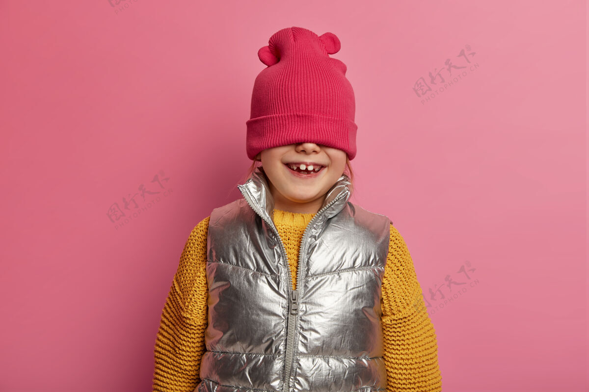 情绪有趣的女孩用帽子遮住脸 积极地咯咯笑 在周围嬉戏 穿着针织的黄色毛衣和背心 在室内对着粉色的墙壁摆姿势 表达快乐的情绪顽皮的孩子在室内欢呼牙牙学语粉彩