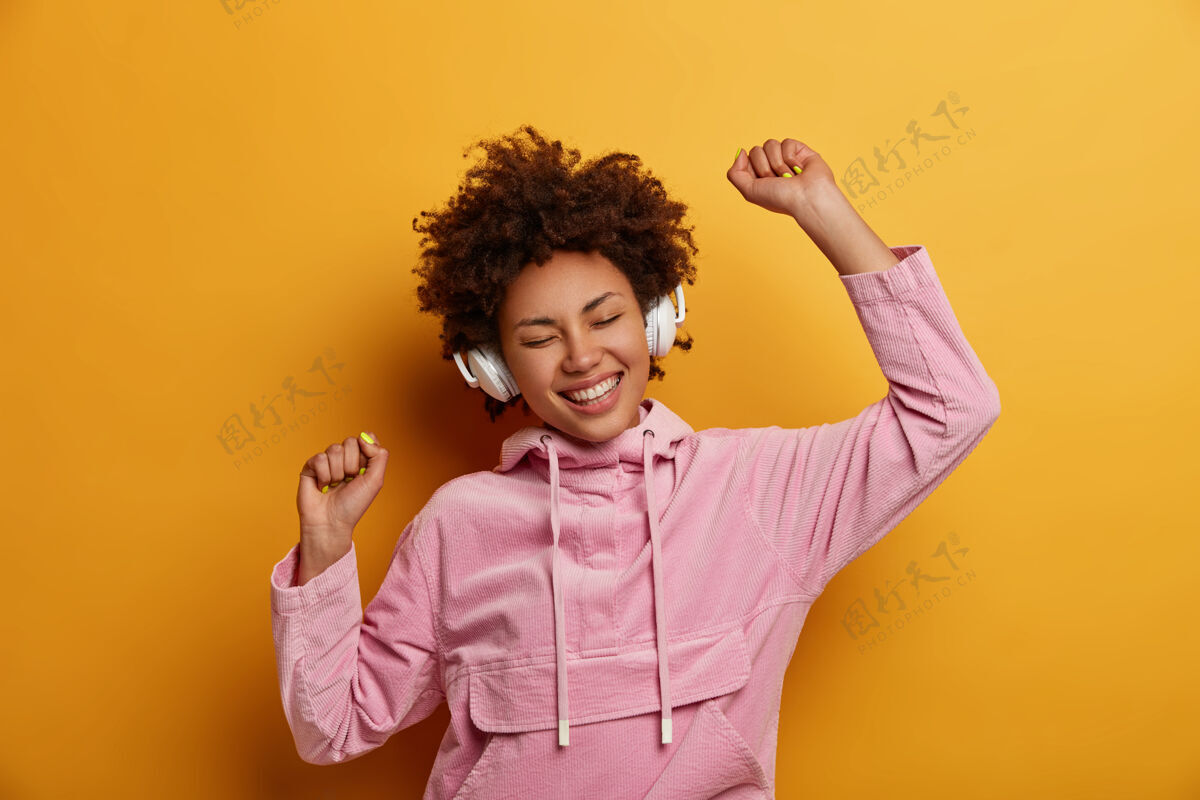 休闲快乐富于表现力的女性戴着耳机听音乐 享受悦耳的旋律 心情愉快 舞姿悠闲 笑容灿烂 穿着粉色运动衫 对着黄色的墙壁摆姿势人 休闲无线耳机快乐