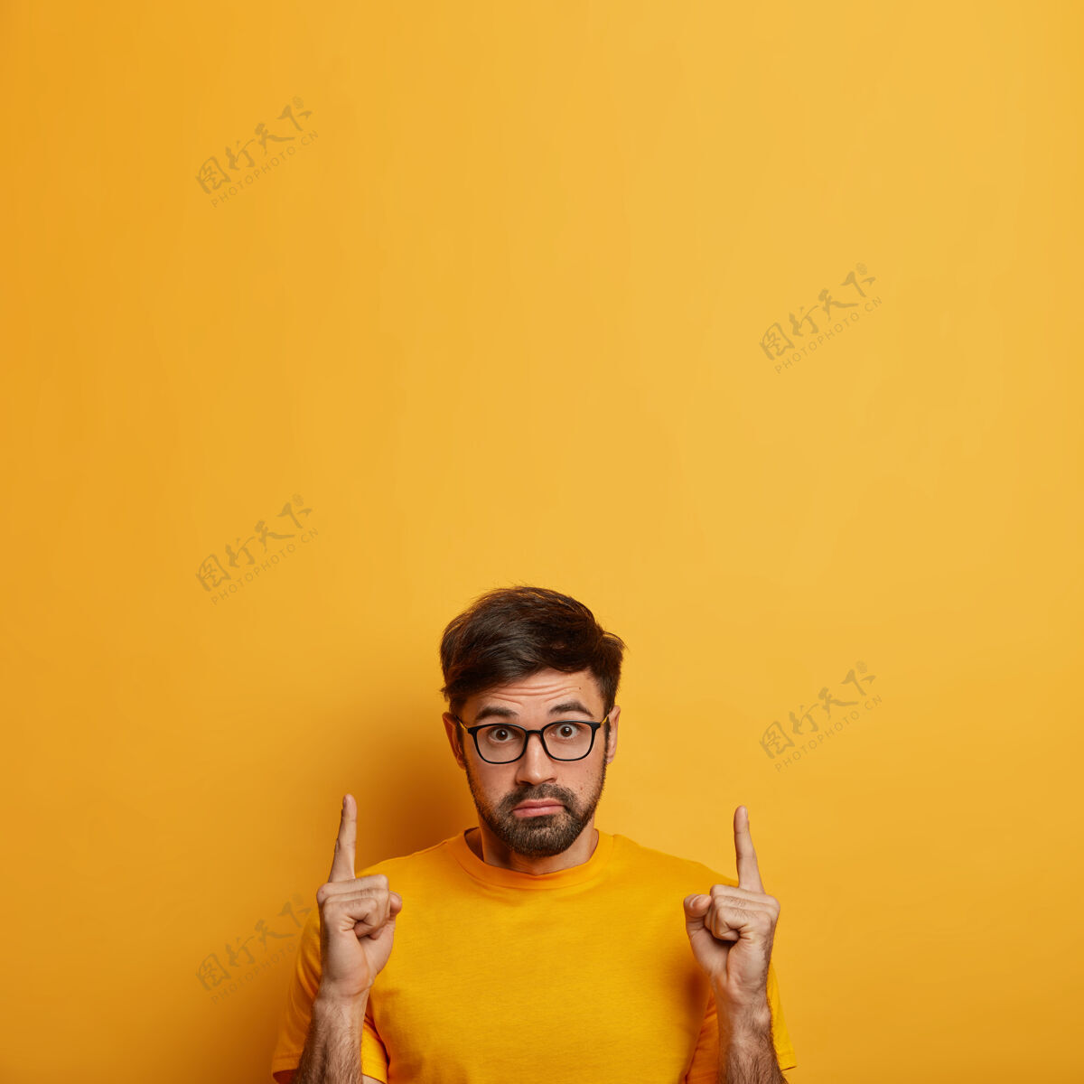 向上惊呆男人的照片展示产品 用惊奇的表情推荐服务 用窃听的眼睛盯着 在空白处向上指示 戴着眼镜 隔离在黄色的墙上模特Omg惊呆