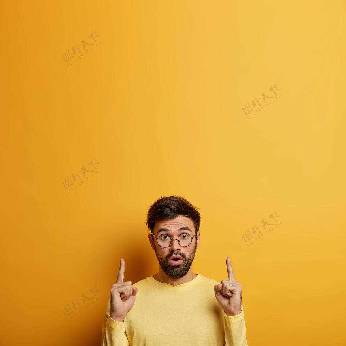 难以置信惊讶的留着胡须的男学生用食指指着上面 演示新产品 讨论销售 因恐惧而喘息 在黄色的墙上摆姿势 为你的促销内容留空广告凝视室内